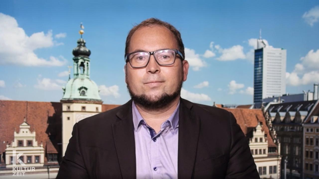 Ein Mann mit Brille und Bart lächelt vor dem Hintergrund einer Stadt und blauem Himmel