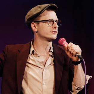 Moritz Neumeier steht in der Mitte des Bildes und hält ein Mikrofon in der Hand. Er blickt in Richtung des rechten Bildrandes.