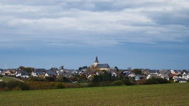 Blick auf ein kleines Dorf mit Kirche hinter einem Feld, auf dem es grün sprießt. Darüber blauer Himmel.