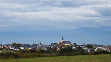 Blick auf ein kleines Dorf mit Kirche hinter einem Feld, auf dem es grün sprießt. Darüber blauer Himmel.