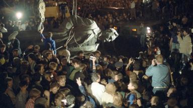 Szene bei Nacht: Eine Menschenmenge schaut zu und fotografiert, wie eine riesige sowjetische Statue von einem Mann mit Seilen vom Sockel heruntergeholt und abtransportiert wird.