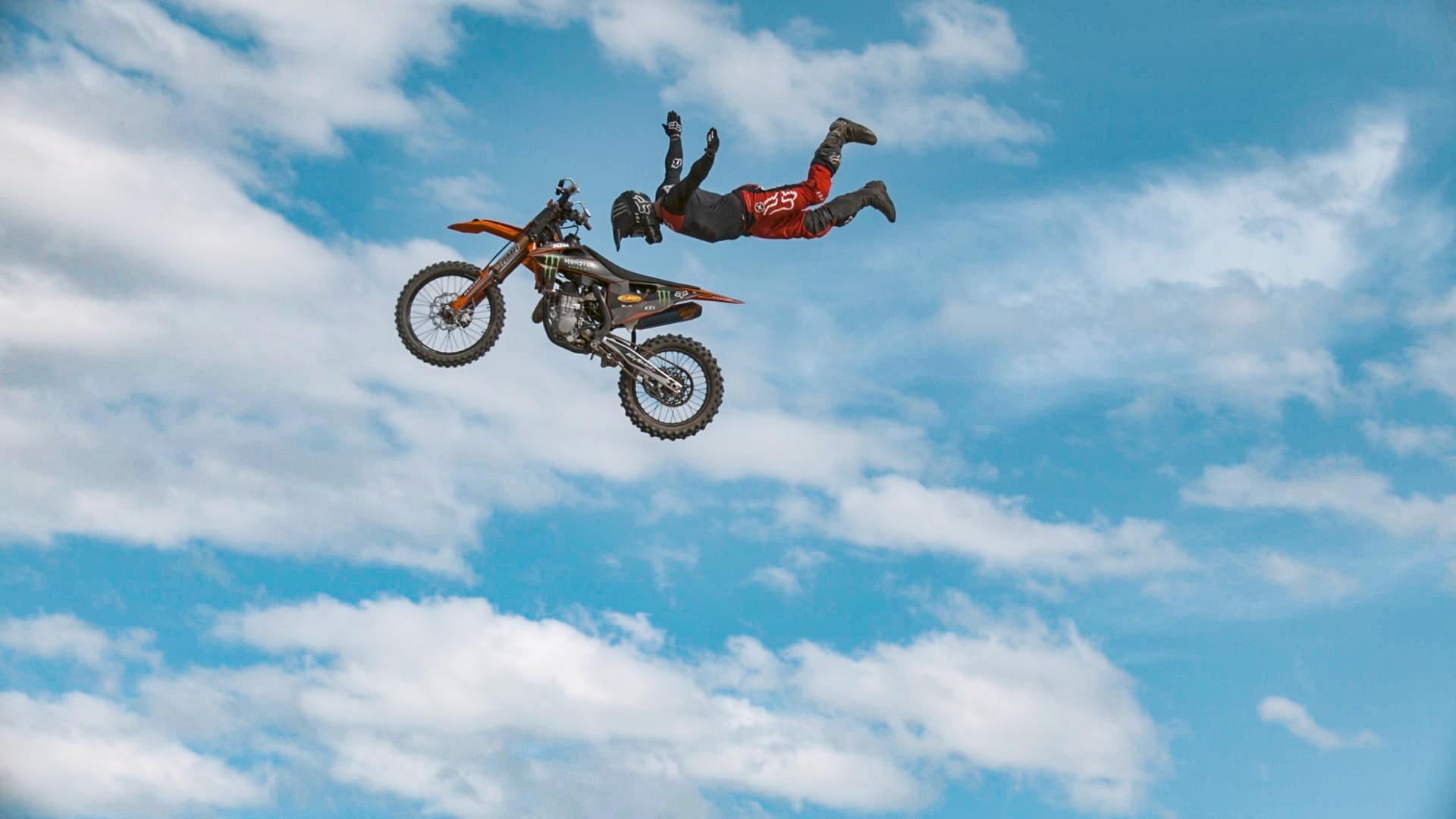 Ein rot-schwarzes Motorrad und ein Stuntman mit schwarzem Helm und roter Hose fliegen durch die Luft vor blauem Himmel mit Wolken. Der Stuntman schwebt über dem Motorrad und hat die Arme nach hinten gestreckt.