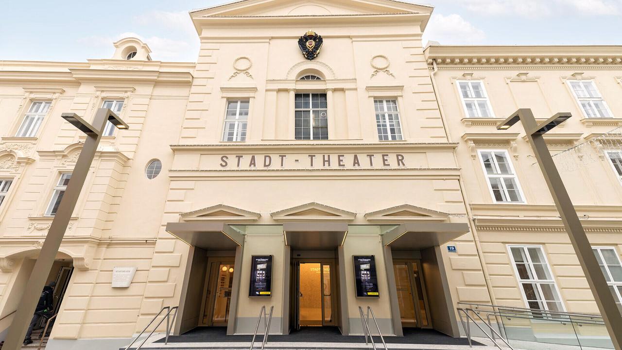 Das Bild zeigt die Fassade des Stadttheaters Wiener Neustadt. Die Architektur des Gebäudes ist klassisch und elegant, mit einem hellen, cremefarbenen Anstrich. Über dem Haupteingang prangt der Name „STADT-THEATER“ in großen, goldenen Buchstaben. 

Die Fassade hat symmetrische Fenster in mehreren Etagen, die den Raum lichtdurchfluten. Ein großes, mittleres Fenster ist von Säulen flankiert, während oben ein Wappen angebracht ist. Vor dem Eingang befinden sich zwei moderne, überdachte Treppenaufgänge, die zu den Glastüren führen. 

Beide Seiten des Eingangs sind mit großformatigen Plakaten ausgestattet, die vermutlich Informationen über Veranstaltungen enthalten. Der Himmel im Hintergrund ist leicht bewölkt und verleiht der Szene eine angenehme Lichtstimmung. 

Insgesamt vermittelt die Aufnahme einen Eindruck von festlicher Atmosphäre, passend zu einem kulturellen Ereignis.