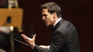 Das Bild zeigt einen männlichen Dirigenten, der sich zur Seite wendet. Er trägt einen schwarzen Frack und hat ein helles Hemd mit Kläppchenkragen an. Sein Gesichtsausdruck ist intensiv und engagiert, während er mit einer Hand gestikuliert. In der anderen Hand hält er einen Dirigentenstab, mit dem er die musikalischen Anweisungen gibt. Im Hintergrund sind unscharfe Konturen von Musikinstrumenten und Personen zu erkennen, die Teil des Orchesters sind. Die Szene vermittelt eine festliche und konzentrierte Atmosphäre, die für ein klassisches Konzert typisch ist.