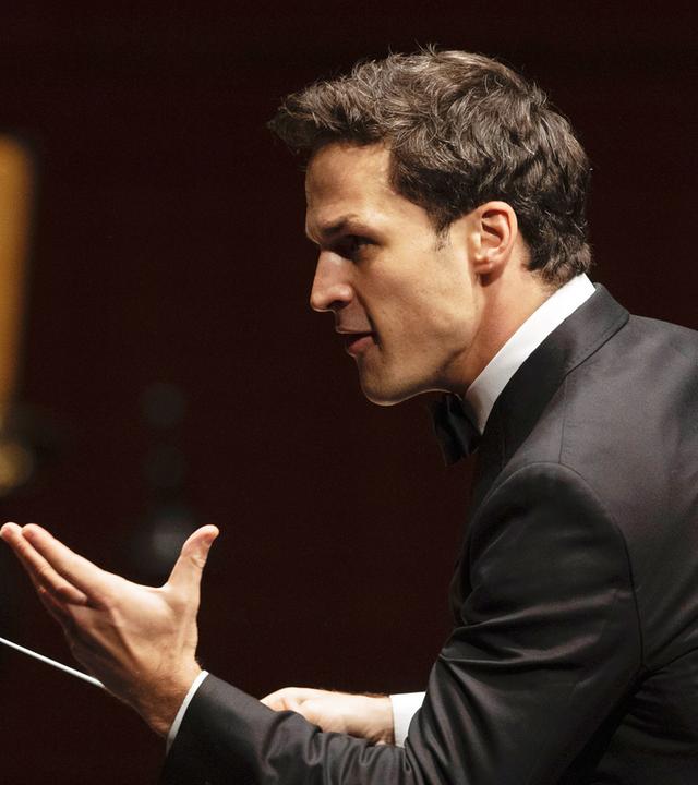 Das Bild zeigt einen männlichen Dirigenten, der sich zur Seite wendet. Er trägt einen schwarzen Frack und hat ein helles Hemd mit Kläppchenkragen an. Sein Gesichtsausdruck ist intensiv und engagiert, während er mit einer Hand gestikuliert. In der anderen Hand hält er einen Dirigentenstab, mit dem er die musikalischen Anweisungen gibt. Im Hintergrund sind unscharfe Konturen von Musikinstrumenten und Personen zu erkennen, die Teil des Orchesters sind. Die Szene vermittelt eine festliche und konzentrierte Atmosphäre, die für ein klassisches Konzert typisch ist.