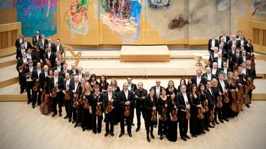 Das Bild zeigt das Mozarteumorchester Salzburg in einem Konzertsaal. Die Orchester-Musiker stehen in mehreren Reihen auf einer hellen Holzfläche. Im Hintergrund ist eine große, künstlerische Wandmalerei zu sehen, die in verschiedenen Farben gestaltet ist und abstrakte Formen zeigt.  Das Orchester besteht aus zahlreichen Musikern, die hauptsächlich in schwarzer Konzertkleidung gekleidet sind. Die Mitglieder halten verschiedene Instrumente, vor allem Geigen. In der Mitte der Gruppe stehen einige Musiker, die im Vordergrund hervorstechen.   Im Saal ist eine reduzierte, aber moderne Bühne erkennbar, die in hellen Holzfarben gehalten ist. Es gibt zudem einige musikalische Instrumente, wie eine Harfe und Schlagzeuge, die im Hintergrund sichtbar sind. Die Beleuchtung des Raumes ist hell und freundlich, wodurch eine einladende Atmosphäre entsteht. Das Bild strahlt eine professionelle und feierliche Stimmung aus, die mit Musik und Kunst verbunden ist.