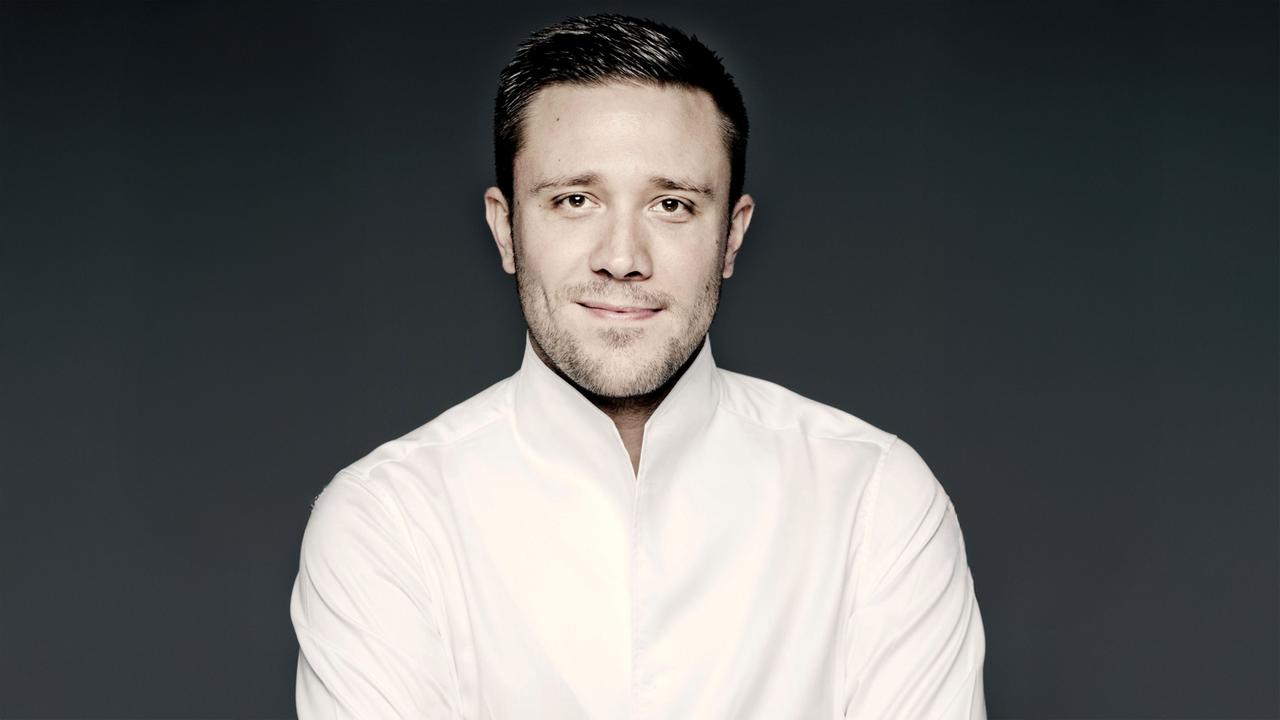 Das Bild zeigt einen Mann mit kurzen, dunklen Haaren und einem leichten Bart. Er trägt ein weißes Hemd mit erhöhtem Kragen, das ihm einen formellen und eleganten Look verleiht. Der Hintergrund ist dunkel, wodurch der Fokus auf seinem Gesicht liegt. Er lächelt leicht und hat einen freundlichen Ausdruck. Der Mann hat braune Augen und eine insgesamt freundliche Ausstrahlung. In der Beschreibung steht, dass es sich um Roberto González-Monjas handelt, der als Dirigent und Geiger tätig ist.