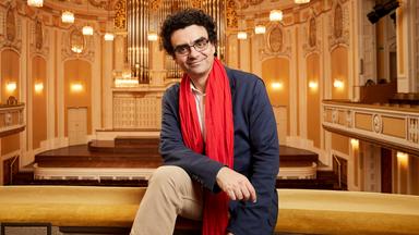 Das Bild zeigt einen Mann, der entspannt auf einer Geländerbrüstung in einem prunkvollen Konzertsaal sitzt. Er trägt ein blaues Sakko, ein helles Hemd und einen auffälligen roten Schal. Der Mann hat lockiges, dunkles Haar und eine Brille.   Im Hintergrund sind die charakteristischen Merkmale des Konzertsaals zu sehen: hohe, mit kunstvollen Verzierungen dekorierte Wände in sanften Farbtönen, mehrere raffinierte Kronleuchter, die den Raum beleuchten, und ein großes, aufwendiges Orgelinstrument an der Wand. Die Bühne ist mit hellem Holz ausgelegt und hat Treppenstufen, die ins Publikum führen. Die gesamte Atmosphäre vermittelt einen eleganten und festlichen Eindruck, passend zu einem Musikfestival.