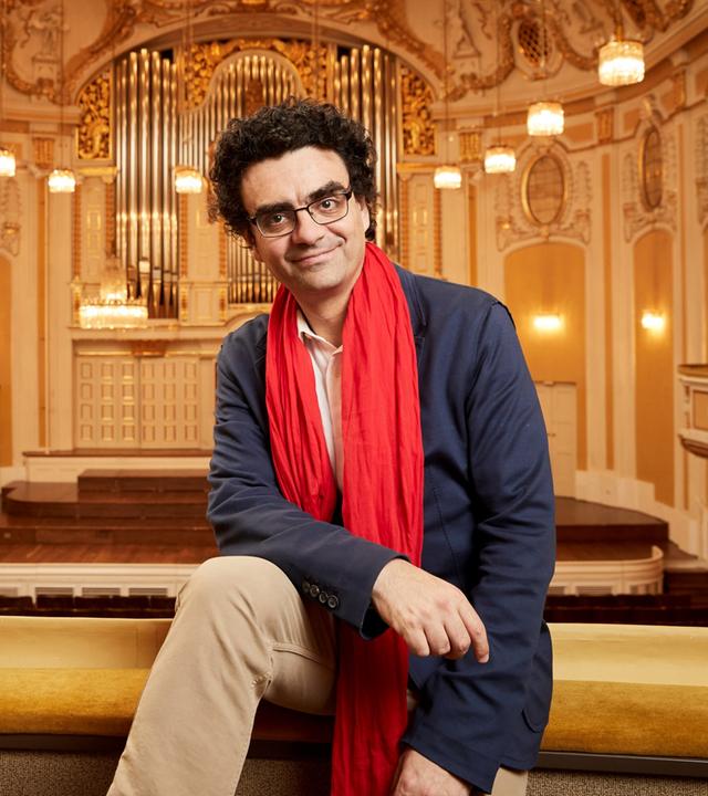 Das Bild zeigt einen Mann, der entspannt auf einer Geländerbrüstung in einem prunkvollen Konzertsaal sitzt. Er trägt ein blaues Sakko, ein helles Hemd und einen auffälligen roten Schal. Der Mann hat lockiges, dunkles Haar und eine Brille.   Im Hintergrund sind die charakteristischen Merkmale des Konzertsaals zu sehen: hohe, mit kunstvollen Verzierungen dekorierte Wände in sanften Farbtönen, mehrere raffinierte Kronleuchter, die den Raum beleuchten, und ein großes, aufwendiges Orgelinstrument an der Wand. Die Bühne ist mit hellem Holz ausgelegt und hat Treppenstufen, die ins Publikum führen. Die gesamte Atmosphäre vermittelt einen eleganten und festlichen Eindruck, passend zu einem Musikfestival.