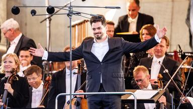 Ein Dirigent steht mit ausgebreiteten Armen vor seinem Orchester und lächelt dem Publikum zu.