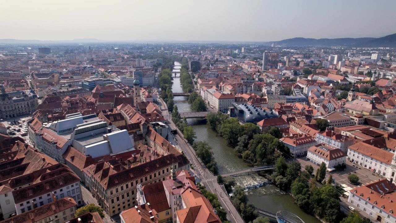 Das Bild zeigt eine Luftaufnahme einer Stadtlandschaft in Österreich, vermutlich Graz. Im Vordergrund sind zahlreiche Dächer von Gebäuden mit roten Ziegeldächern zu sehen. Einige der Gebäude haben unterschiedliche Höhen und Architekturen, wobei einige modern und andere eher traditionell wirken.  Durch die Stadt fließt der Fluss Mur, der von grünen Uferbereichen gesäumt ist. Auf dem Fluss sind eventuell einige kleine Boote zu erkennen. Es gibt mehrere Brücken, die über den Fluss führen. Im Hintergrund sind weitere Stadtgebäude sichtbar, darunter hohe Bauwerke und Wohnblocks sowie hügelige Landschaften am Horizont.  Das Bild hat eine klare Atmosphäre und wirkt hell, was auf sonniges Wetter hindeutet. Die gesamte Szene vermittelt das Gefühl einer lebendigen Stadt mit einer Mischung aus Natur und urbanen Strukturen.