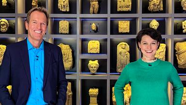 Moderator mit weiblichem Gast vor Wand mit Exponaten.