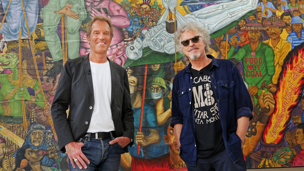 Der Moderator und sein Gast stehen vor einem gemalten Bild mit Demonstranten.