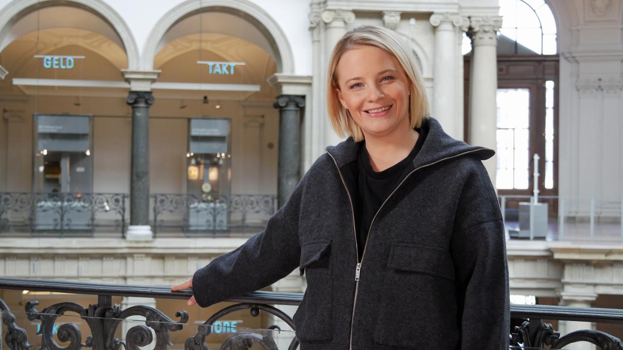 Eine blonde Frau in grauer Jacke steht auf einem Balkon im Museum. Gegenüber sind Torbögen und gusseiserne, verzierte Geländer zu sehen.