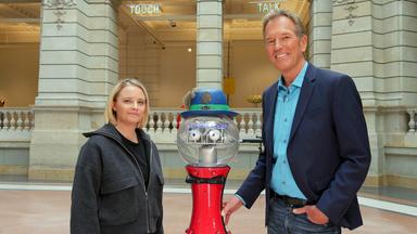 Jasna Fritzi Bauer und Markus Brock stehen rechts und links eines Roboters mit rotem Fuß und durchsichtigem Glaskopf mit Hut.