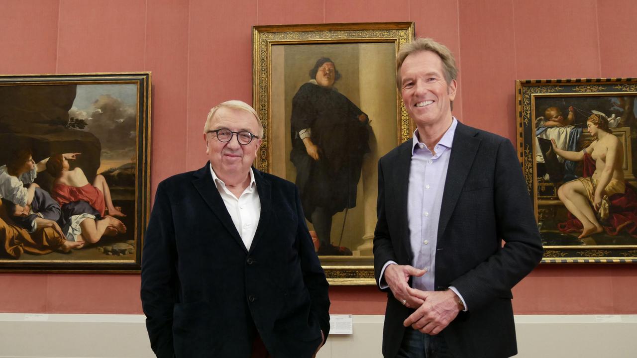 Der Moderator und der Buchautor vor einer roten Wand mit drei Gemälden in Goldrahmen