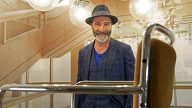 Ein bärtiger Mann mit Hut hinter einem Stuhl. Über ihm mehrere Glaskugel-Lampen