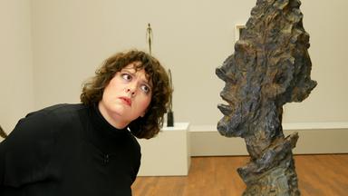 Frau im dunklen Pulli beugt sich mit fragendem Gesicht zu einer Metallbüste