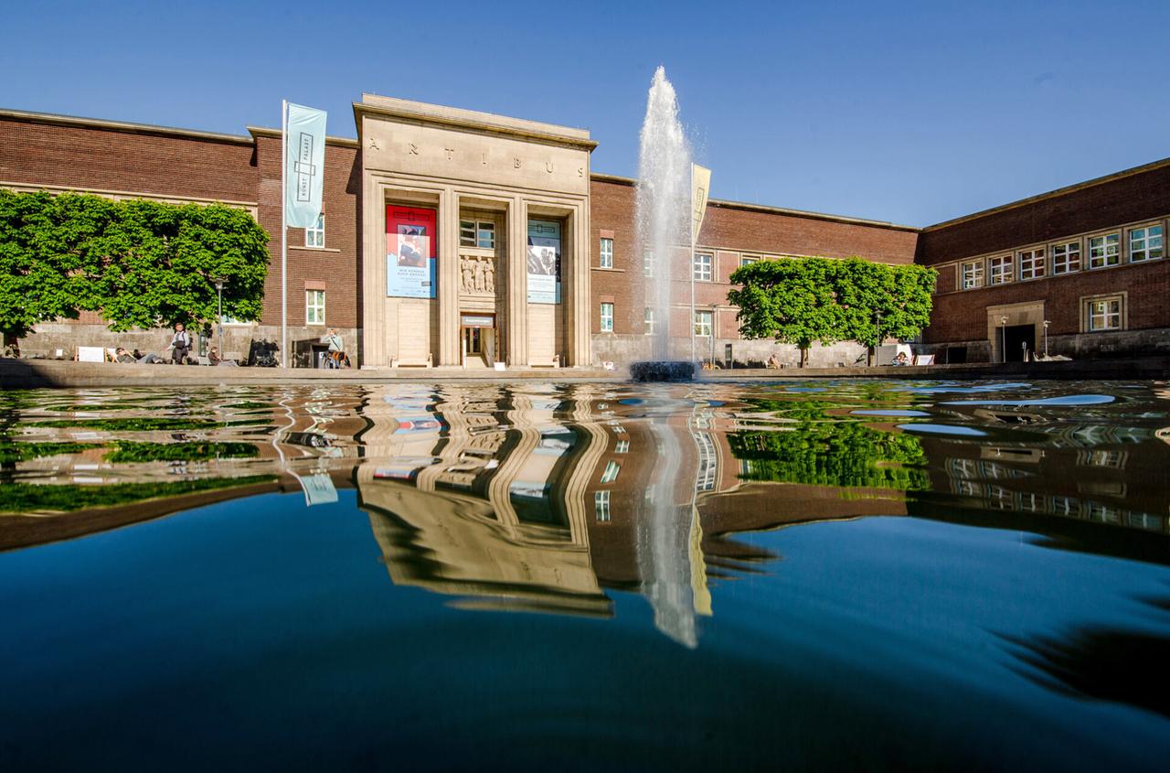 Ein Gebäude und ein Springbrunnen spiegeln sich in einem Gewässer