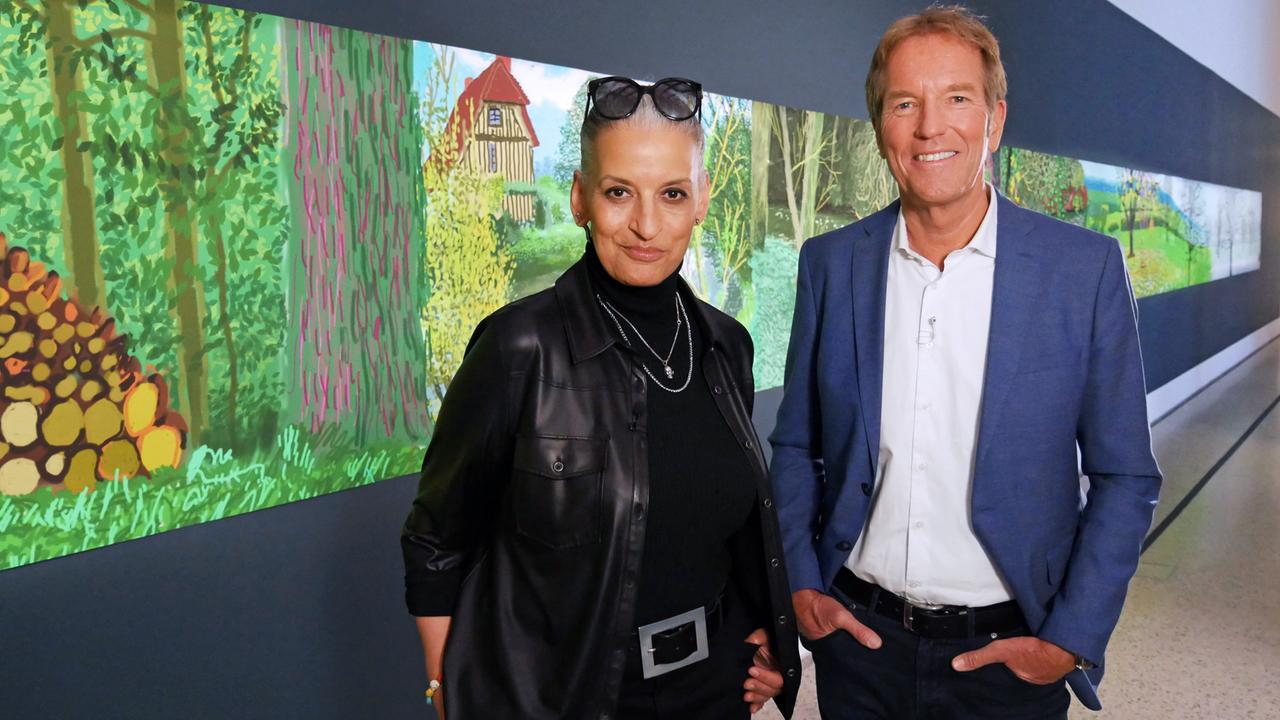 Eine Frau und ein Mann stehen vor einem langen gemalten Bild einer idyllischen Landschaft mit Haus.