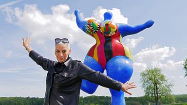 Eine Frau posiert lächelnd vor einer riesigen blauen weiblichen Statue mit bunten Brüsten.