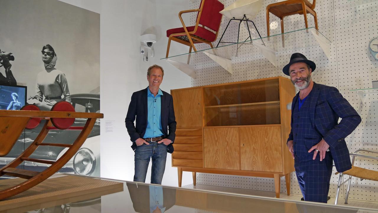 Der Moderator und der Gast stehen zwischen Stühlen und einer Kommode im Museum.