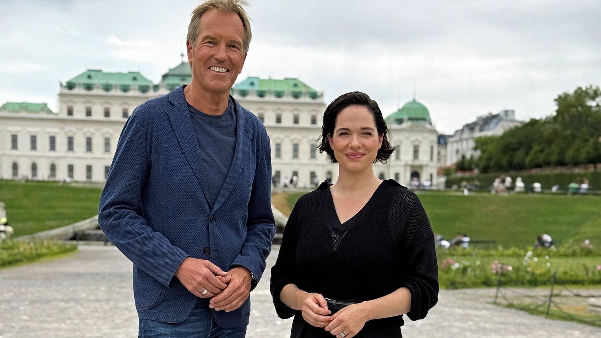 Markus Brock steht lächelnd mit Verena Altenberger im Park vor historischen weißen Gebäude mit grünem Dach.