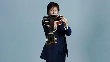 Paul McCartney hält seine Gitarre Richtung Kamera.