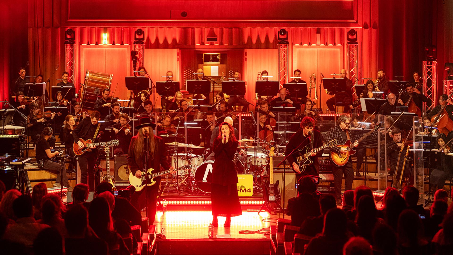 Man sieht einen beleuchteten Konzertsaal mit einer Band, einer Sängerin, einem Orchester und Publikum.