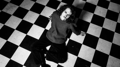 Auf einer schwarz-weiß-Fotografie sieht man einen jungen Mann der auf einem schwarz-weiß-gefliestem Boden sitzt.