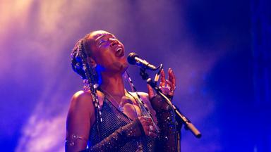 Eine schwarze Frau in glitzerndem Kostüm singt auf einer Bühne.