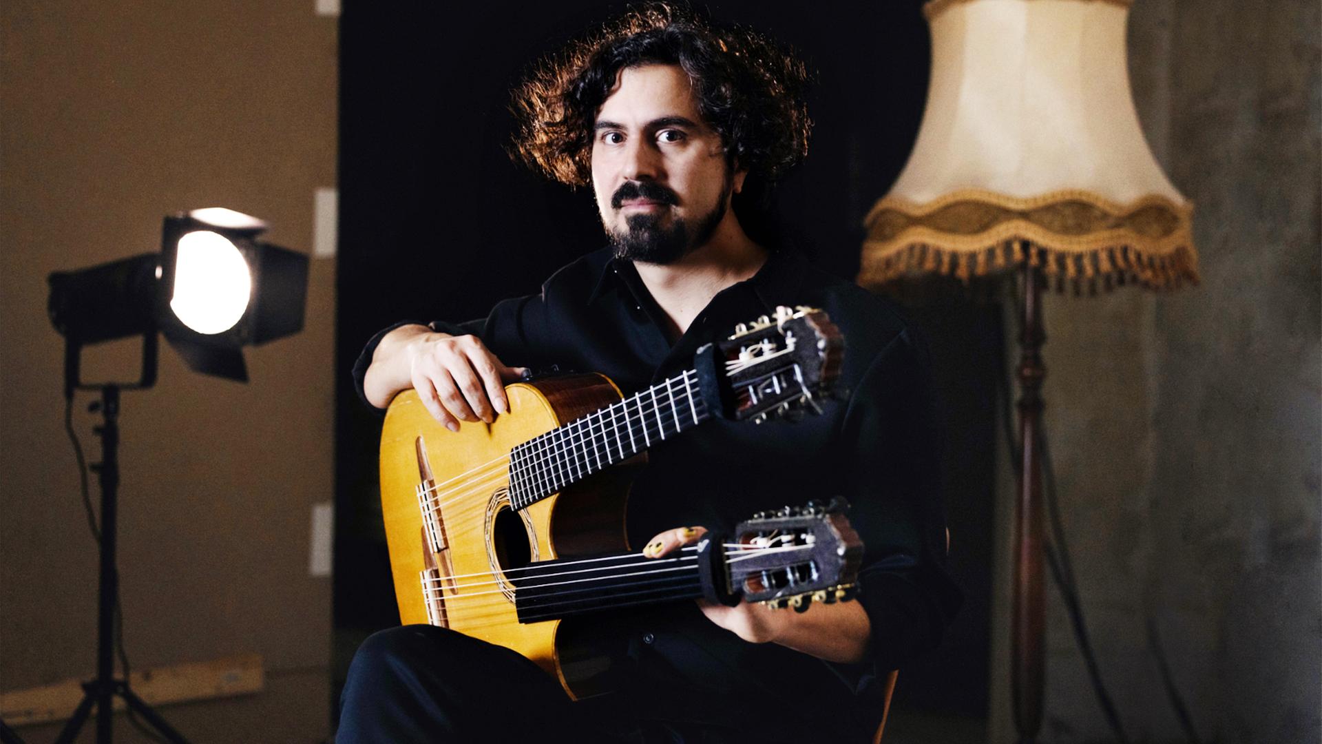 Ein Mann mit einer Doppelhals- Gitarre sitzt zwischen einem Scheinwerfer und einer Stehlampe.