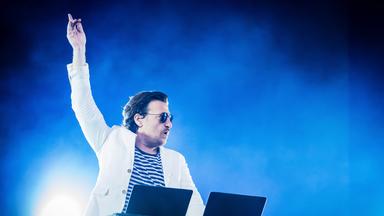 Ein Mann, der einen weißen Anzug trägt, streckt den Arm empor. Er steht hinter einem DJ-Pult auf einer Bühne.