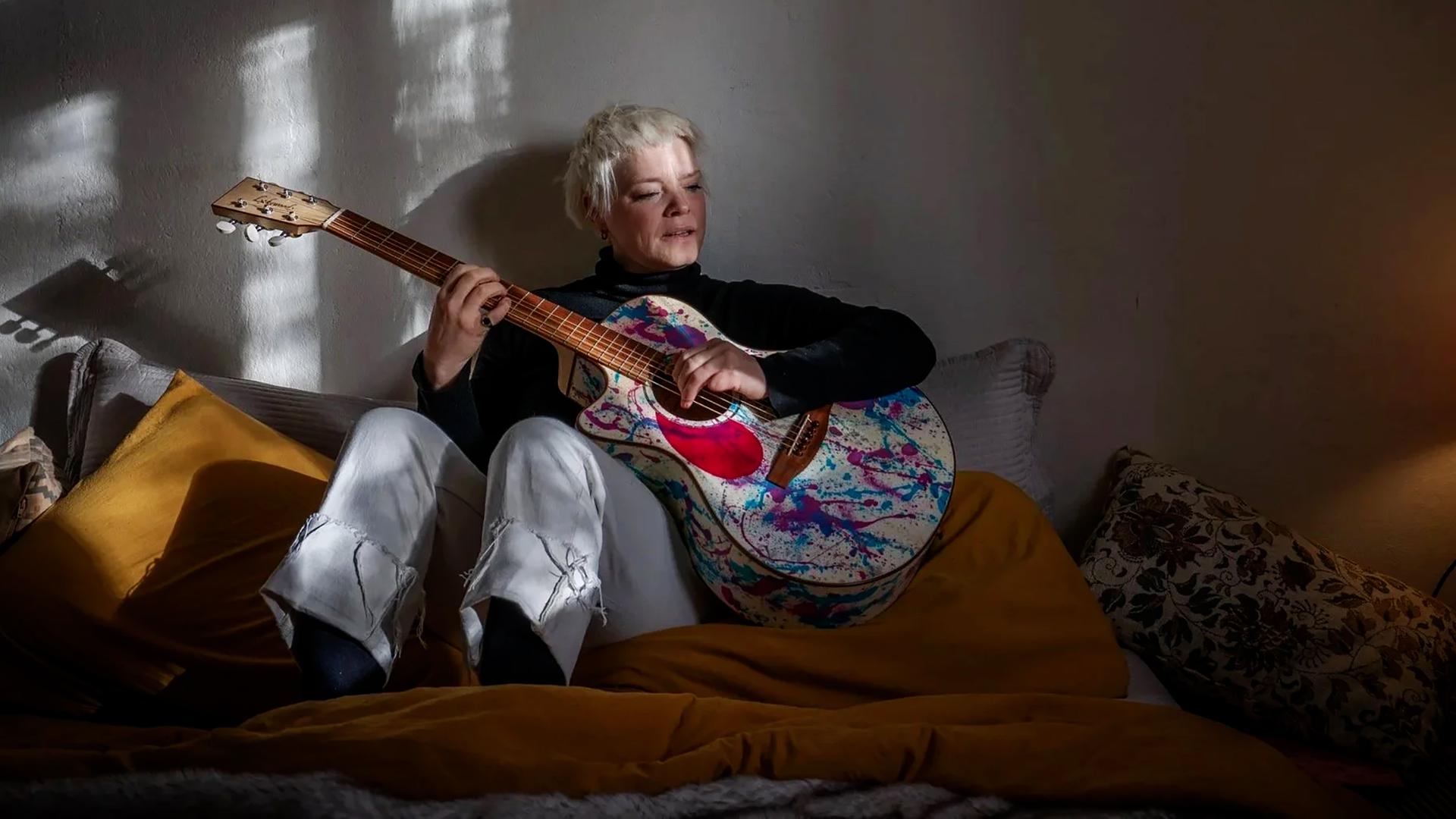 Eine blonde Frau sitzt in einem abgedunkelten Raum auf einem Bett und spielt Gitarre.