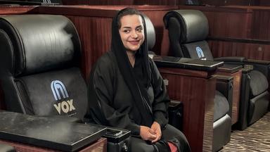 Eine lächelnde Frau sitzt in einem großen schwarzen Kinositz, sie trägt einen schwarzen Mantel und ein schwarzes Tuch über den Haaren.