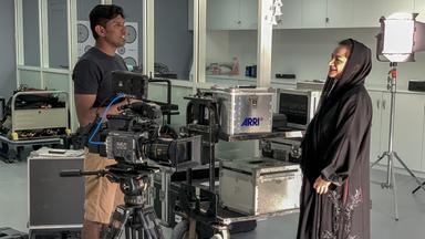 Rechts im Bild steht eine Frau in schwarzer Kleidung mit schwarzem Tuch über den Haaren, links ein Mann in kurzer Hose und T-Shirt. Sie stehen in Mitten von Kamera-Equipment, im Vordergrund sieht man eine Fernsehkamera auf einem Stativ. 