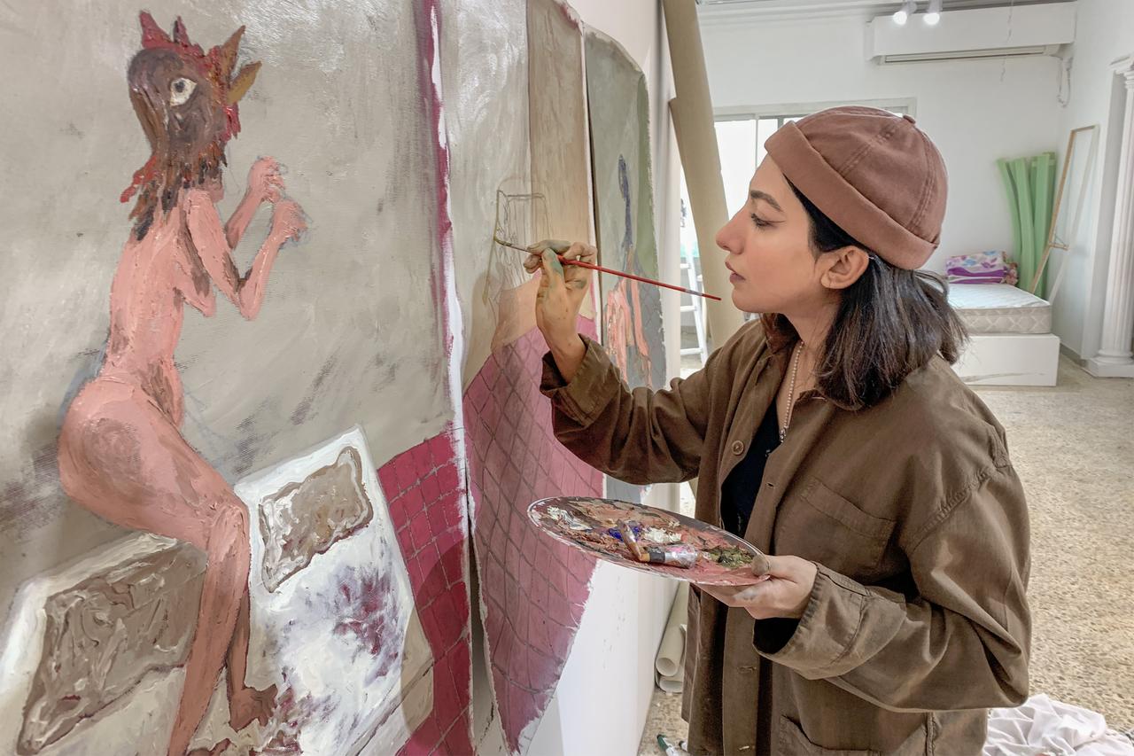 Man sieht eine junge, dunkelhaarige Frau seitlich vor einer bemalten Leinwand, sie hält eine Farbpalette und einen Pinsel in der Hand und malt an dem Bild.