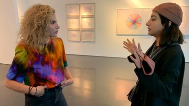 Man sieht zwei Frauen seitlich, die sich unterhalten. Die eine trägt ein buntes Oberteil und hört zu, die andere trägt eine Mütze und gestikuliert. Die beiden stehen in einer Galerie. 