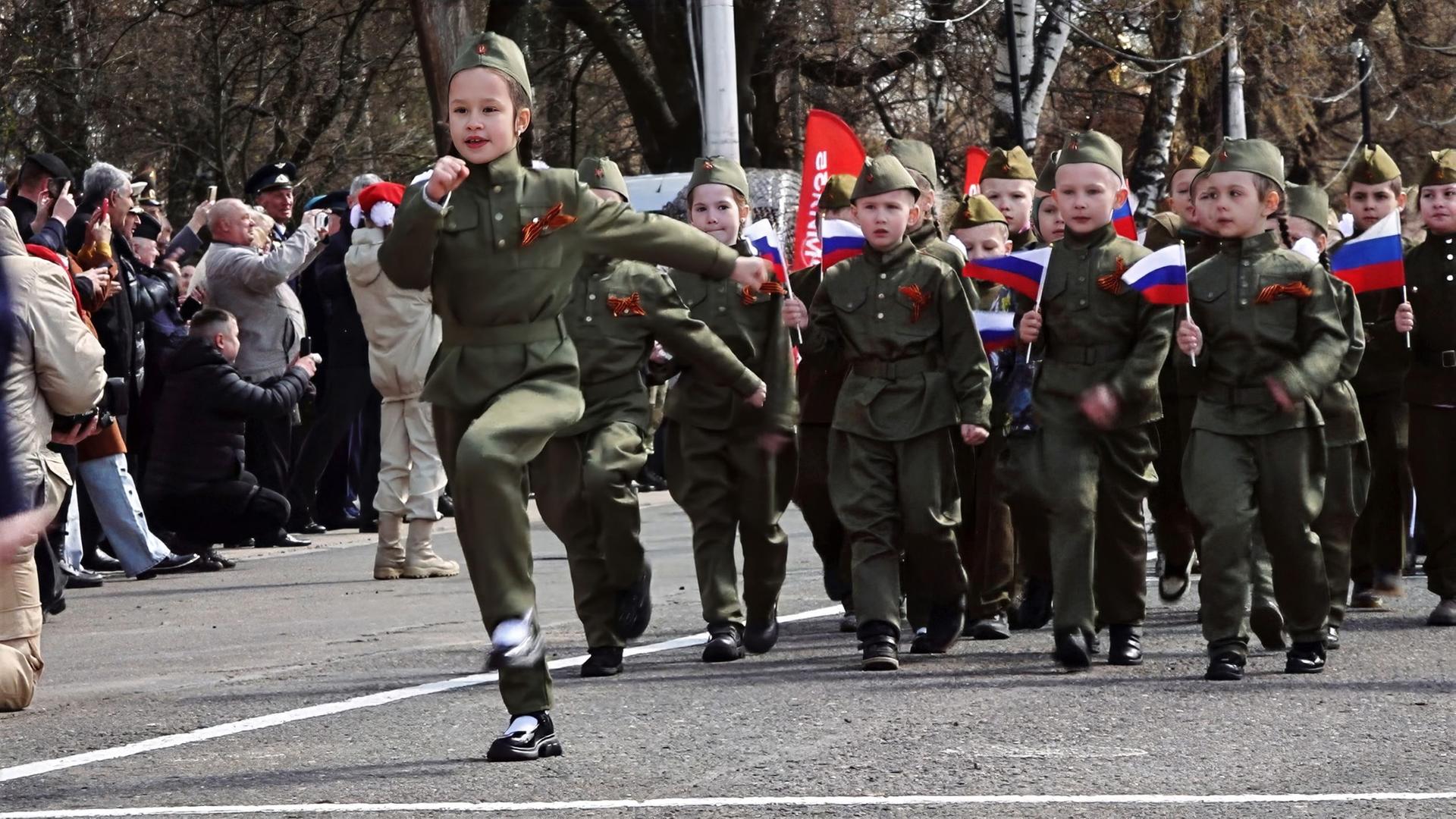 Kinder in Militäruniformen marschieren bei einer Parade, einige tragen russische Fahnen, im Hintergrund Publikum mit Kameras.