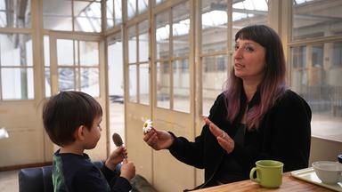 Eine Frau mit dunklem, schulterlangem Haar spricht mit einem kleinen Jungen in einem lichtdurchfluteten Raum. Die Frau hält eine Gänseblümchenblüte in der Hand, während der Junge einen braunen Samenstand betrachtet. Auf dem Tisch stehen eine grüne Tasse und ein weißer Becher.