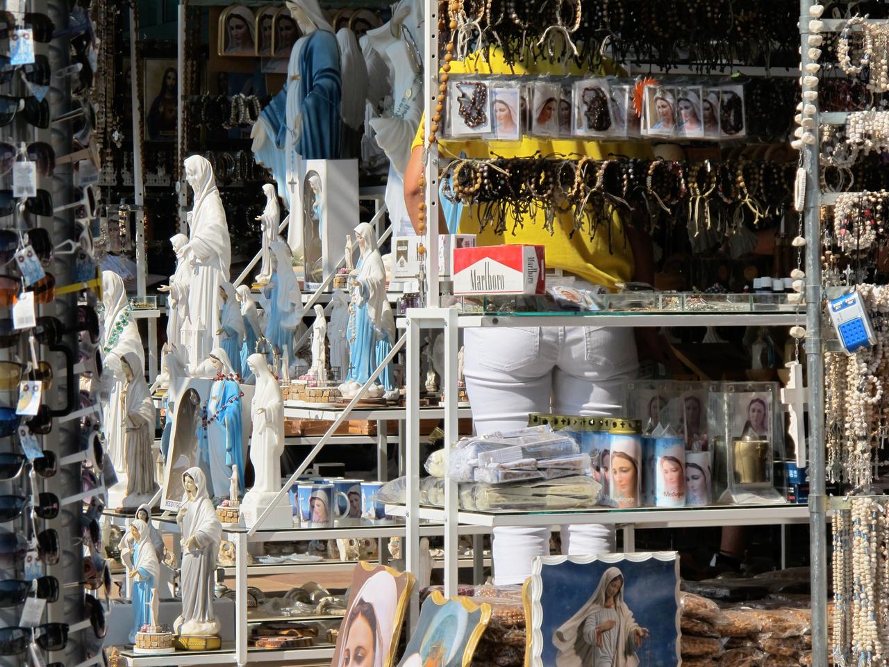 Das Bild zeigt einen Souvenirladen an einem Wallfahrtsort, vermutlich in Medjugorje. Im Vordergrund sind zahlreiche Figuren und Statuen der Jungfrau Maria auf Regalen ausgestellt. Diese Statuen haben meist weiße Gewänder und viele sind mit blauen Akzenten verziert.   Hinter den Statuen befinden sich verschiedene religiöse Souvenirs, darunter Rosenkränze und Postkarten mit Bildern der Jungfrau Maria. Auf der rechten Seite des Bildes sind einige Statuen in einer klaren Glasvitrine zu sehen, während links eine Stange mit Sonnenbrillen sichtbar ist.   Im Hintergrund ist eine Person in gelber Kleidung erkennbar, die den Rücken zur Kamera hat. Ihre Unterkörperbekleidung besteht aus weißen Hosen. Das gesamte Bild vermittelt eine geschäftige, pilgerartige Atmosphäre, die durch die Vielzahl an religiösen Erinnerungsstücken geprägt ist.