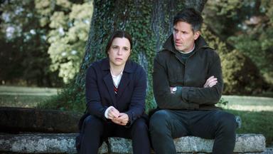 Pia Sander (Annika Kuhl) und Oliver von Bodenstein (Tim Bergmann) sitzen im Halbdunkel auf einem großen Stein unter einem großen Baumstamm. Pia stützt ihre Arme auf den Knien ab und blickt angespannt in die Ferne, Oliver schaut zu seiner Kollegin. Er hat die Arme verschränkt.