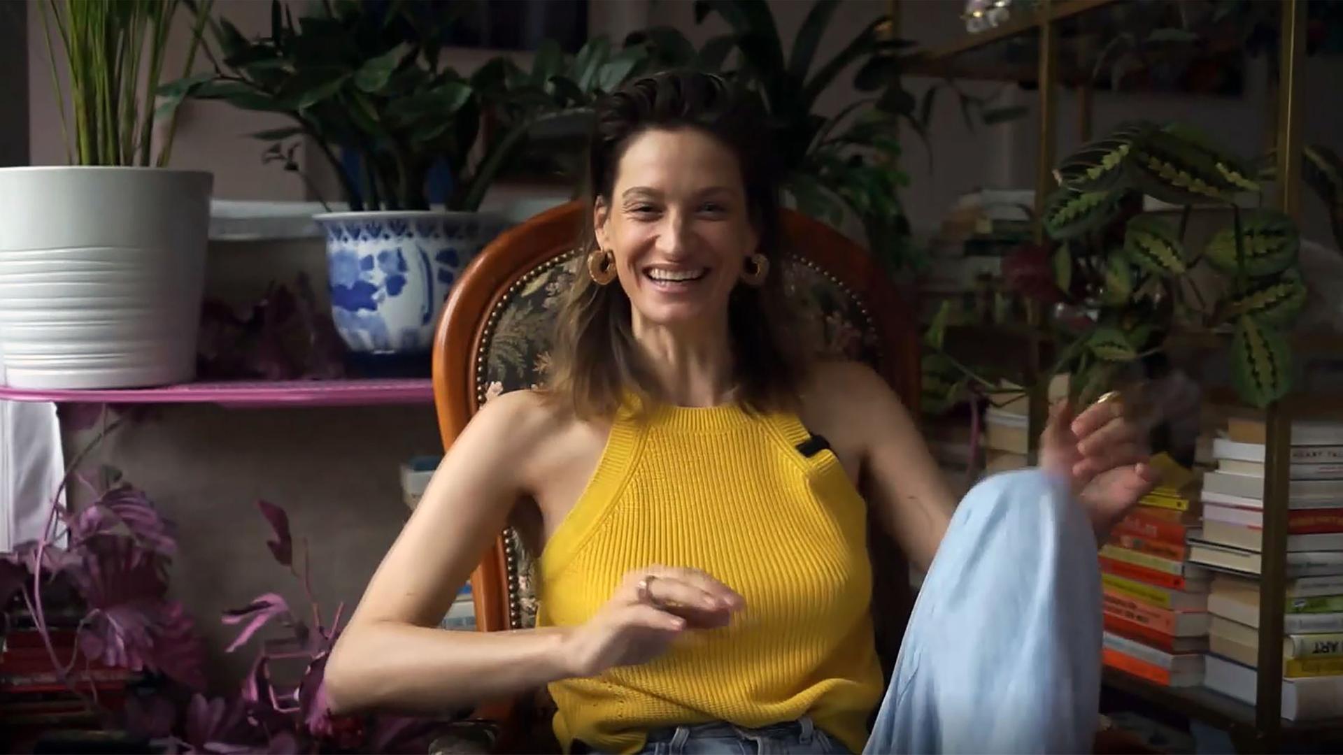 Annique Delphine sitzt auf einem gepolsterten Stuhl. Ihr linkes Bein ist angezogen und sie lacht. Im Hintergrund und auf der rechten Seite sind offene, halbhohe Regale mit Grünpflanzen und Büchern zu sehen. 