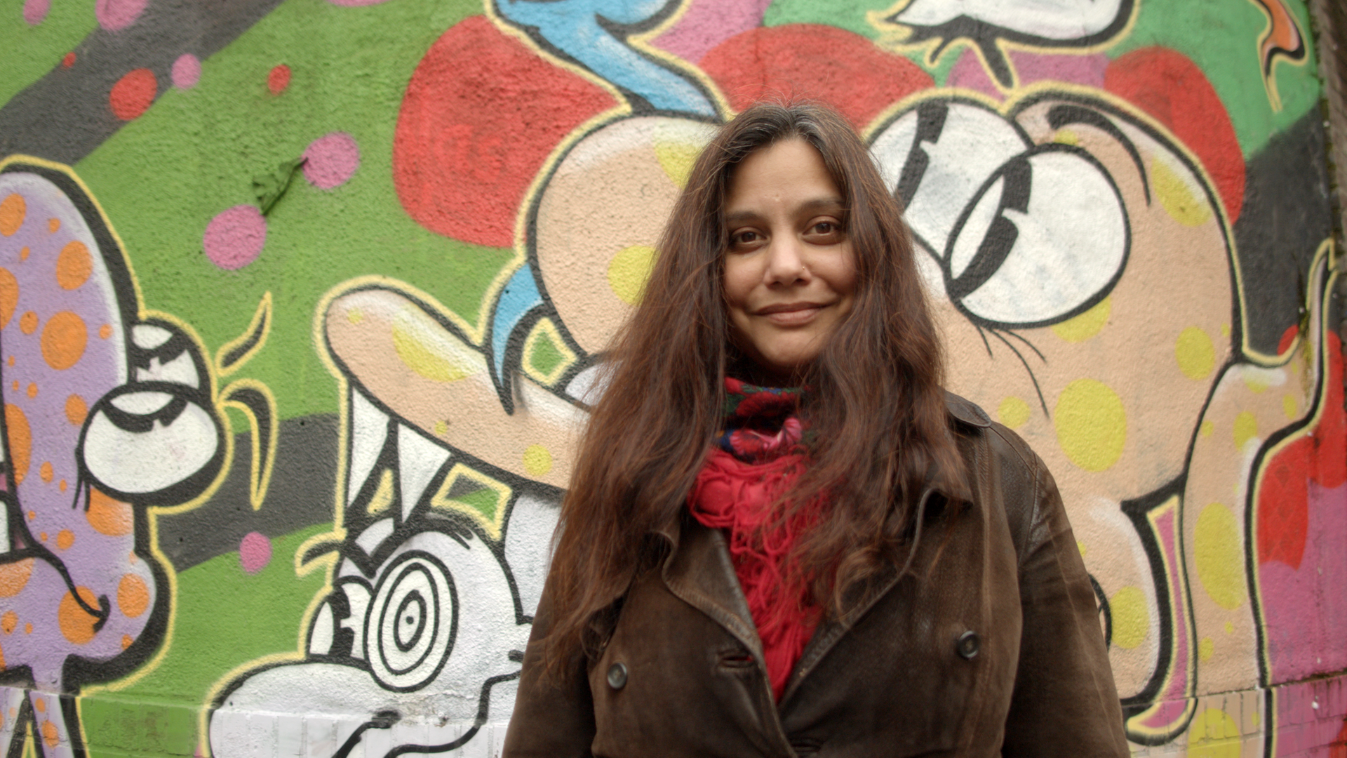 Die Journalistin und Kulturwissenschaftlerin Mithu Sanyal steht vor einer bunten Graffiti-Wand in Düsseldorf und lächelt in die Kamera. Sie hat lange braune Haare und trägt einen brauen Mantel.