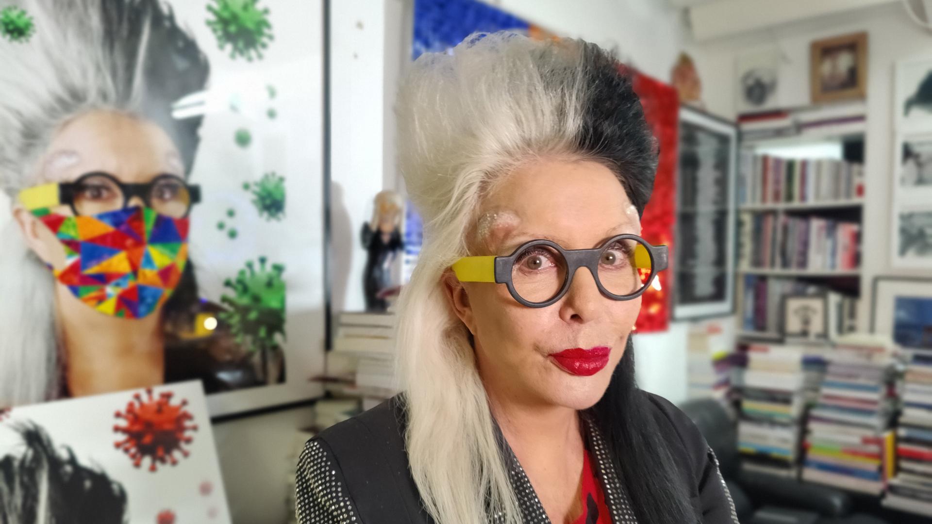 Die Performance- und Multimedia-Künstlerin ORLAN in ihrem Atelier in Paris. Sie hat schwarz-weiße Haare, trägt eine gelb-schwarze Brille und schaut zur Seite. An ihrer Stirn kann man Narben erkennen. Im Hintergrund sind zwei Selbstportraits von ihr zu sehen.