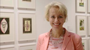 Kuratorin Dr. Gabriele Schor steht in einem Ausstellungraum, hinter ihr hängen Bilder an der Wand. Sie hat blonde Haare, trägt eine Kette, einen lachsfarbenen Blazer und lacht in die Kamera.