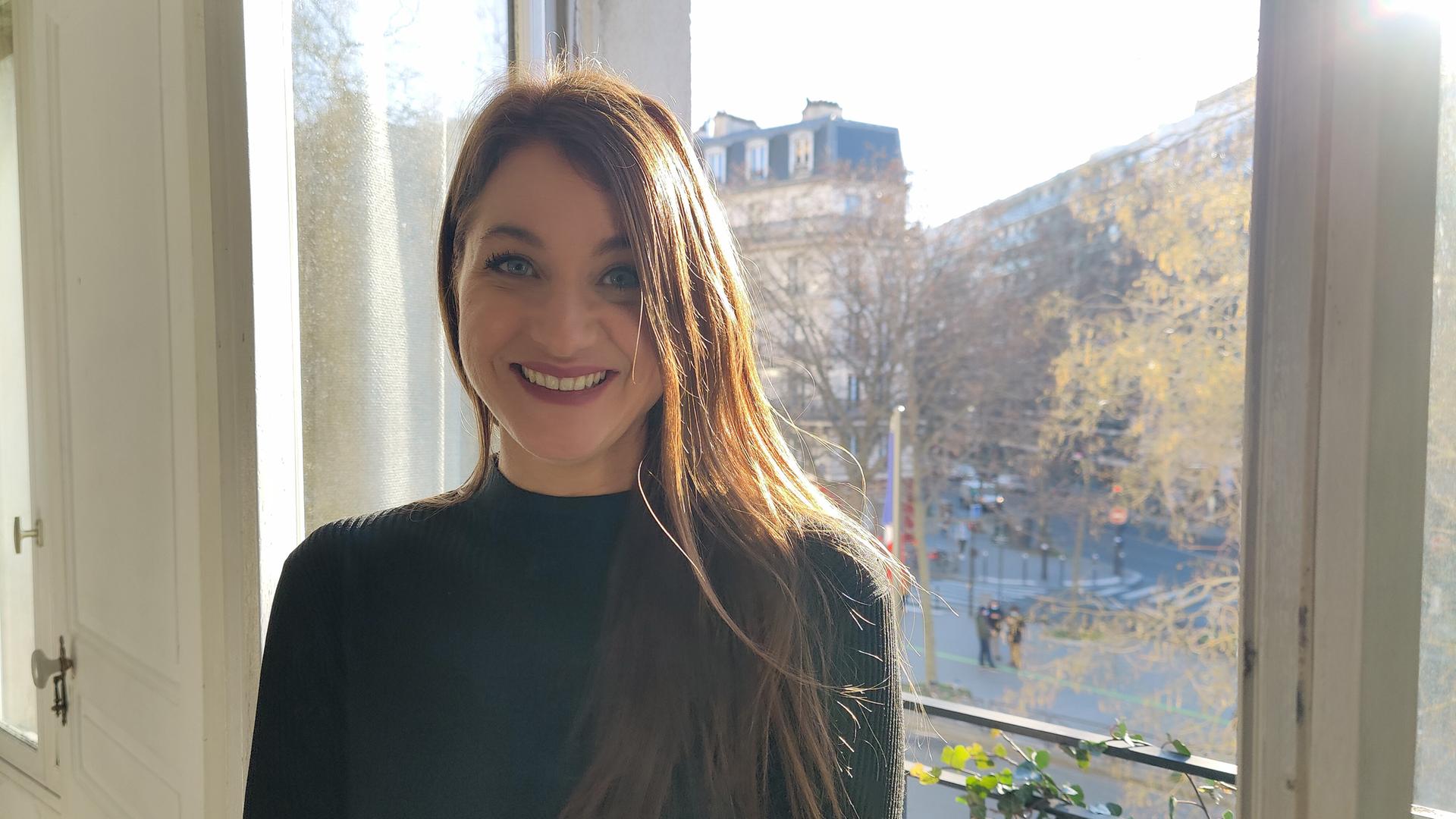 Die Performance-Künstlerin Deborah de Robertis steht vor einer Terrassentür in einer Wohnung in Paris. Im Hintergrund sind eine breite Straße, kahle Bäume und mehrgeschossige Wohnbebauung zu sehen. Es scheint die Sonne.