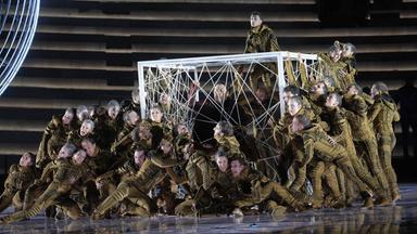 Nahaufnahme Bühne mit vielen Akteuren in grünen Anzügen um weißen Drahtwürfel herum