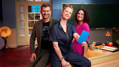 Zwei Frauen und ein Mann sitzen auf einem Lehrerpult und lächeln in die Kamera.