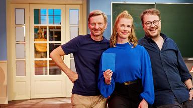 Eine Frau und zwei Männer stehen lächelnd vor einer Tafel und einer Fenstertür im Studio.